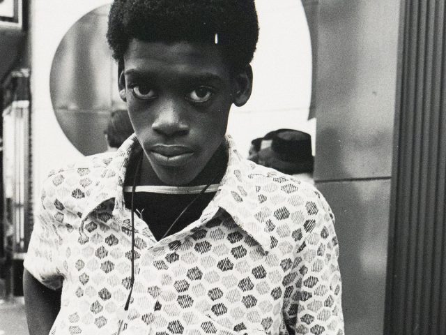 A black and white portrait shows a young boy with short, dark curly hair, leaning against a bicycle. He wears a patterned, short-sleeved shirt, light-colored pants, and a key on a string around his neck, gazing directly at the viewer with a serious expression.