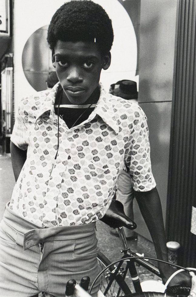 A black and white portrait shows a young boy with short, dark curly hair, leaning against a bicycle. He wears a patterned, short-sleeved shirt, light-colored pants, and a key on a string around his neck, gazing directly at the viewer with a serious expression.