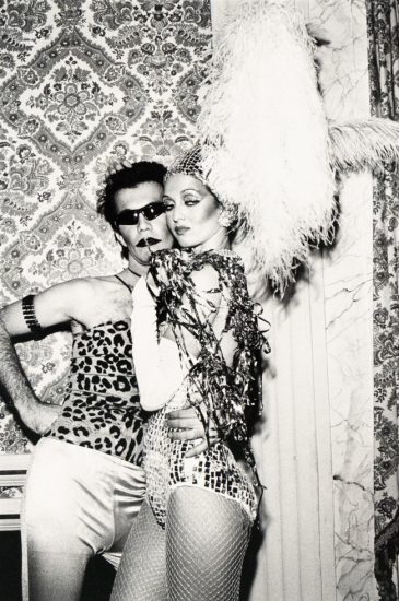 A black and white photograph captures two costumed individuals posing in front of a patterned wallpaper. The person on the left wears sunglasses and a leopard print top, while the person on the right wears a sequined outfit, fishnet stockings, and a large feathery headpiece.