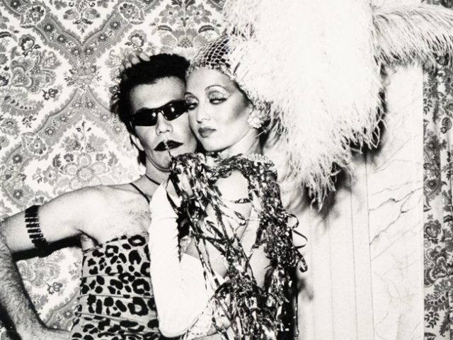 A black and white photograph captures two costumed individuals posing in front of a patterned wallpaper. The person on the left wears sunglasses and a leopard print top, while the person on the right wears a sequined outfit, fishnet stockings, and a large feathery headpiece.