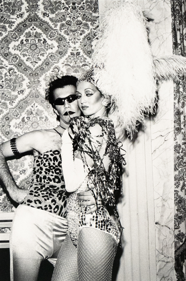 A black and white photograph captures two costumed individuals posing in front of a patterned wallpaper. The person on the left wears sunglasses and a leopard print top, while the person on the right wears a sequined outfit, fishnet stockings, and a large feathery headpiece.