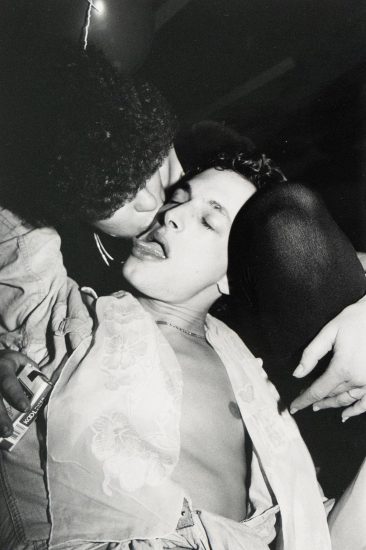 A close-up black and white photograph captures two individuals in an intimate moment, one appearing to kiss the other who has their eyes closed and tongue slightly extended.