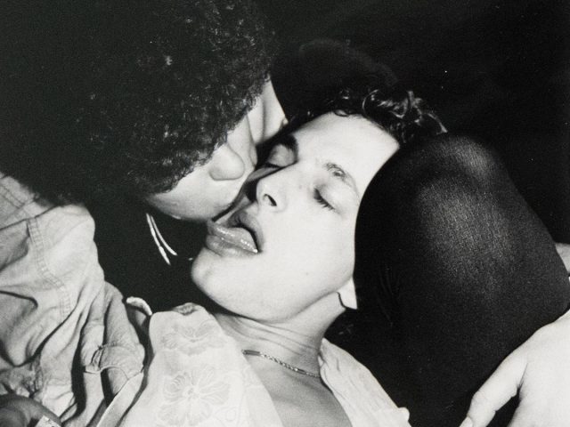 A close-up black and white photograph captures two individuals in an intimate moment, one appearing to kiss the other who has their eyes closed and tongue slightly extended.