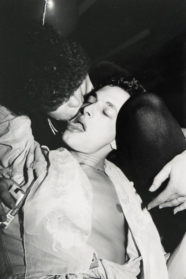 A close-up black and white photograph captures two individuals in an intimate moment, one appearing to kiss the other who has their eyes closed and tongue slightly extended.