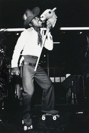 A black and white photograph captures a person dressed in a cowboy hat and outfit, on roller skates, holding a toy stick horse to their face. They are looking upwards and to the right, in what appears to be an indoor setting with dark surroundings.