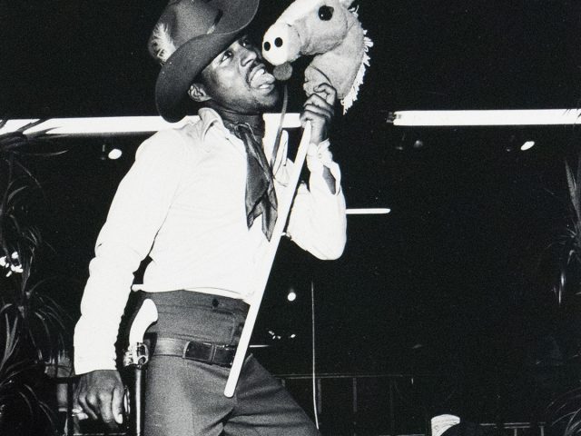 A black and white photograph captures a person dressed in a cowboy hat and outfit, on roller skates, holding a toy stick horse to their face. They are looking upwards and to the right, in what appears to be an indoor setting with dark surroundings.