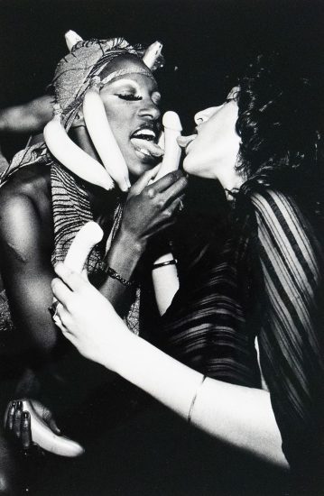 Black-and-white photograph of two people mid-performance or party scene. One person wears a banana headdress and holds a phallic-shaped object near their mouth, while the other leans in with their tongue out. They are surrounded by additional bananas held in various hands, with expressions suggesting playful or theatrical eroticism.