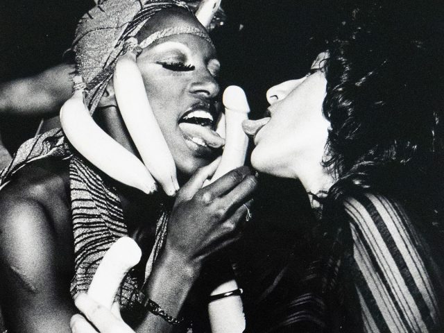 Black-and-white photograph of two people mid-performance or party scene. One person wears a banana headdress and holds a phallic-shaped object near their mouth, while the other leans in with their tongue out. They are surrounded by additional bananas held in various hands, with expressions suggesting playful or theatrical eroticism.