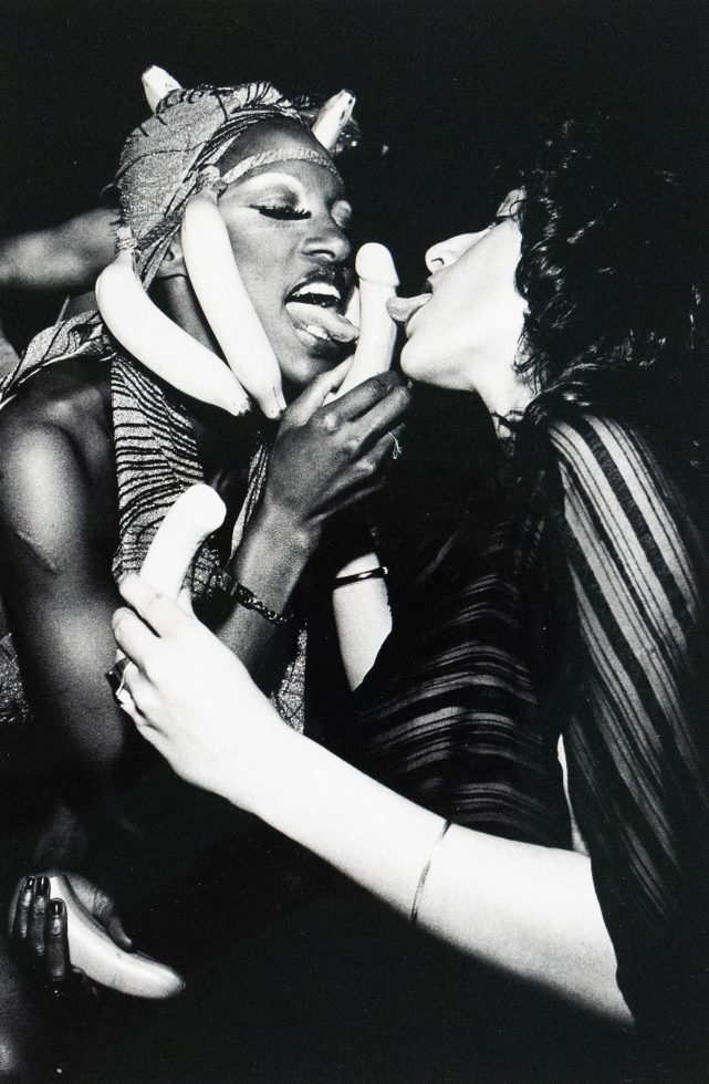 Black-and-white photograph of two people mid-performance or party scene. One person wears a banana headdress and holds a phallic-shaped object near their mouth, while the other leans in with their tongue out. They are surrounded by additional bananas held in various hands, with expressions suggesting playful or theatrical eroticism.