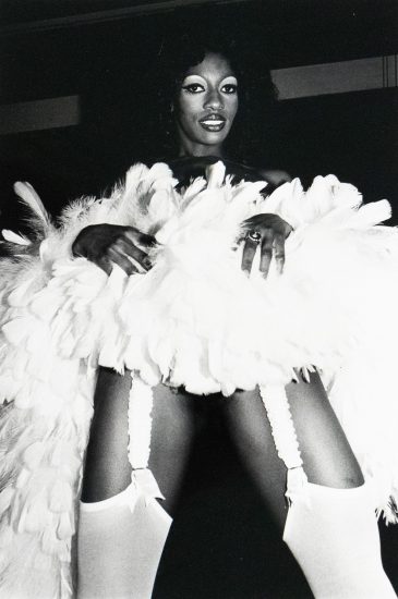 A black and white photograph of a woman with dark hair and striking eye makeup posing in a voluminous white feathered garment with visible stocking garters. She is wearing a large ring and looking directly at the camera.