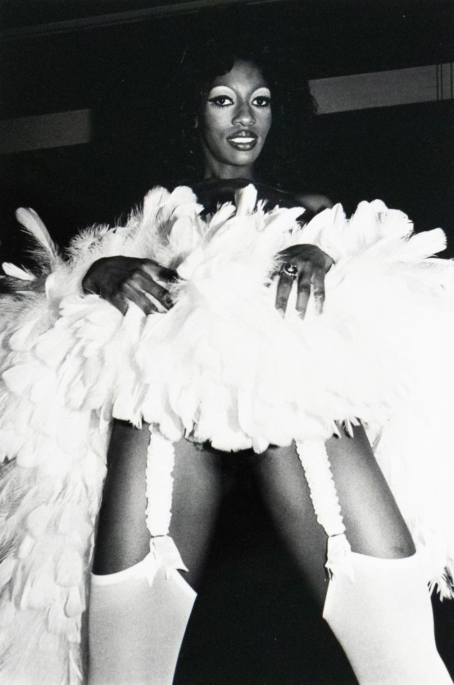 A black and white photograph of a woman with dark hair and striking eye makeup posing in a voluminous white feathered garment with visible stocking garters. She is wearing a large ring and looking directly at the camera.