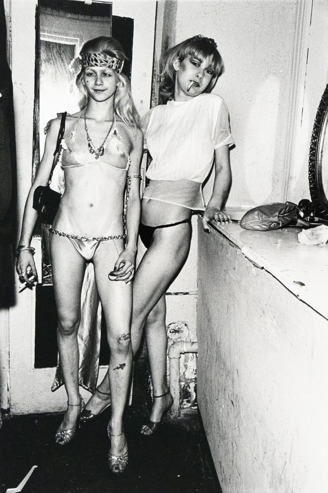 Two women pose in a dimly lit interior; one wears a metallic bikini with a headband and the other a sheer top with underwear. Both hold cigarettes and stand near a scuffed wall and mirror.