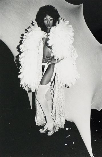 A person with dark, voluminous hair stands in a dramatic pose, wearing a feathered stole, lingerie, stockings, and metallic shoes. The black and white photograph features a stark contrast between the figure and the abstract, light-colored backdrop.