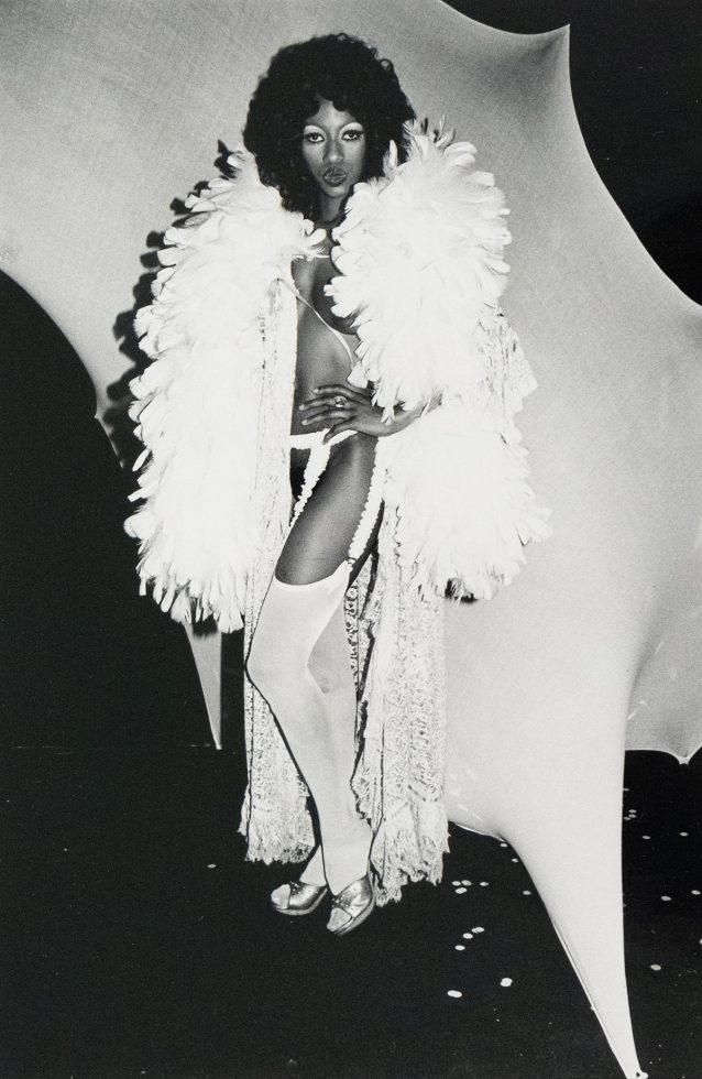 A person with dark, voluminous hair stands in a dramatic pose, wearing a feathered stole, lingerie, stockings, and metallic shoes. The black and white photograph features a stark contrast between the figure and the abstract, light-colored backdrop.