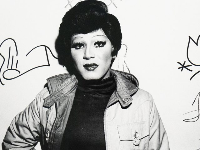 A black and white close-up photograph captures a person identified as a drag queen, with dramatic eye makeup, dark lipstick, and a styled wig, wearing an open jacket over a dark top. They are looking directly at the viewer, and what appear to be signatures or doodles are visible on the white wall behind them.