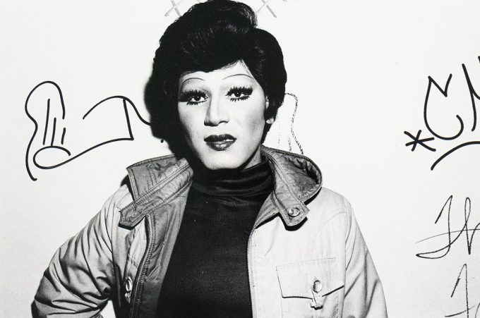 A black and white close-up photograph captures a person identified as a drag queen, with dramatic eye makeup, dark lipstick, and a styled wig, wearing an open jacket over a dark top. They are looking directly at the viewer, and what appear to be signatures or doodles are visible on the white wall behind them.