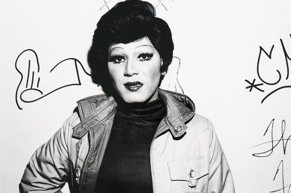 A black and white close-up photograph captures a person identified as a drag queen, with dramatic eye makeup, dark lipstick, and a styled wig, wearing an open jacket over a dark top. They are looking directly at the viewer, and what appear to be signatures or doodles are visible on the white wall behind them.