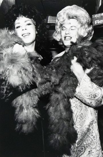 Black-and-white photograph of two people in elaborate wigs and patterned clothing, smiling and holding large feather boas.