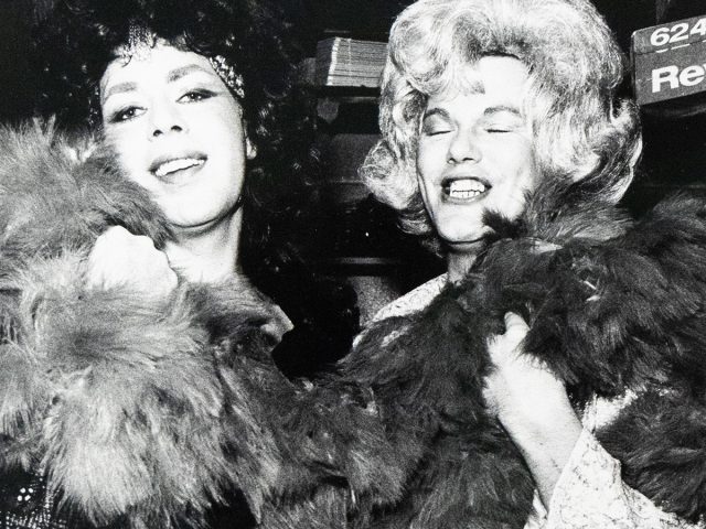 Black-and-white photograph of two people in elaborate wigs and patterned clothing, smiling and holding large feather boas.