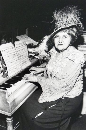 A vintage black and white photograph captures an elderly woman in an elaborate feathered hat, seated at a piano with sheet music. She is depicted playing the instrument and looking towards the viewer.