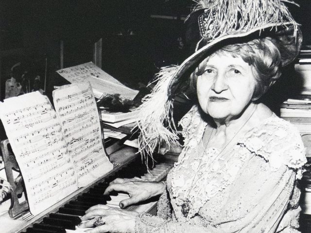 A vintage black and white photograph captures an elderly woman in an elaborate feathered hat, seated at a piano with sheet music. She is depicted playing the instrument and looking towards the viewer.