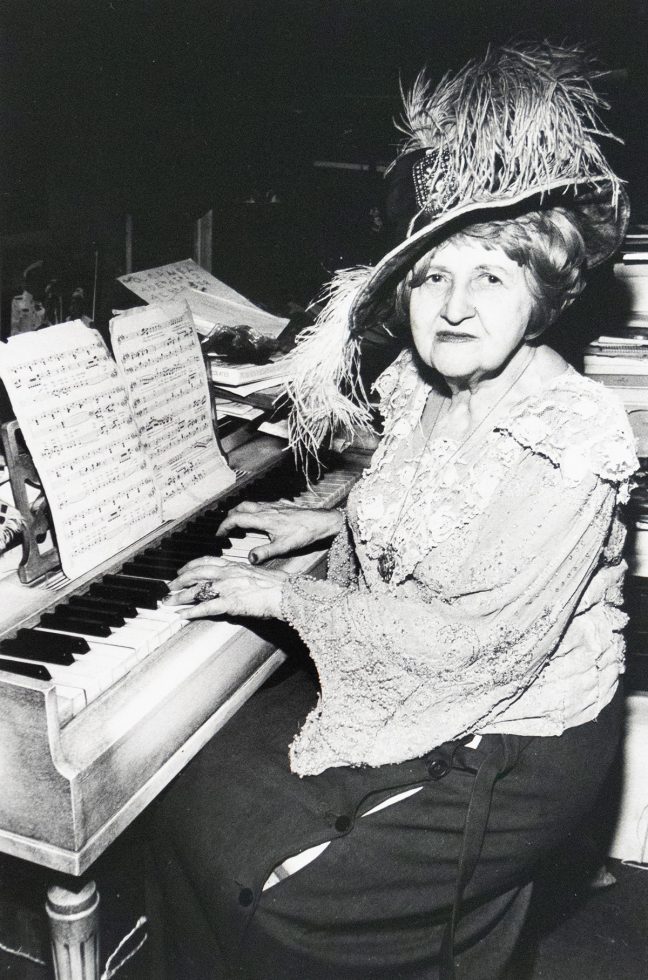 A vintage black and white photograph captures an elderly woman in an elaborate feathered hat, seated at a piano with sheet music. She is depicted playing the instrument and looking towards the viewer.