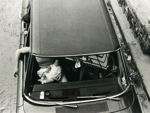 A black and white overhead photograph captures the interior of a car with two adults and an infant. One adult leans out the driver's side window, and the infant is seated in the front passenger area, while the other adult is partially visible in the front passenger seat.