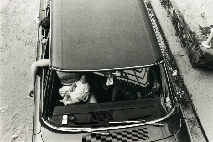 A black and white overhead photograph captures the interior of a car with two adults and an infant. One adult leans out the driver's side window, and the infant is seated in the front passenger area, while the other adult is partially visible in the front passenger seat.