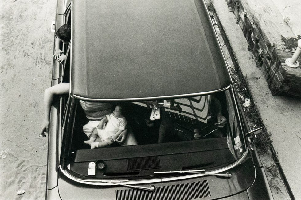 A black and white overhead photograph captures the interior of a car with two adults and an infant. One adult leans out the driver's side window, and the infant is seated in the front passenger area, while the other adult is partially visible in the front passenger seat.