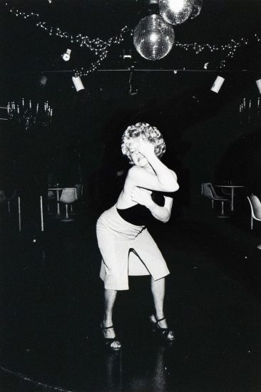 A woman with curly blonde hair and a dark top dances in a dimly lit room, with her hand raised to her face. Disco balls and string lights are visible overhead, suggesting a nightclub or performance venue.