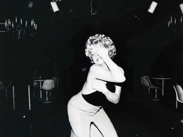 A woman with curly blonde hair and a dark top dances in a dimly lit room, with her hand raised to her face. Disco balls and string lights are visible overhead, suggesting a nightclub or performance venue.