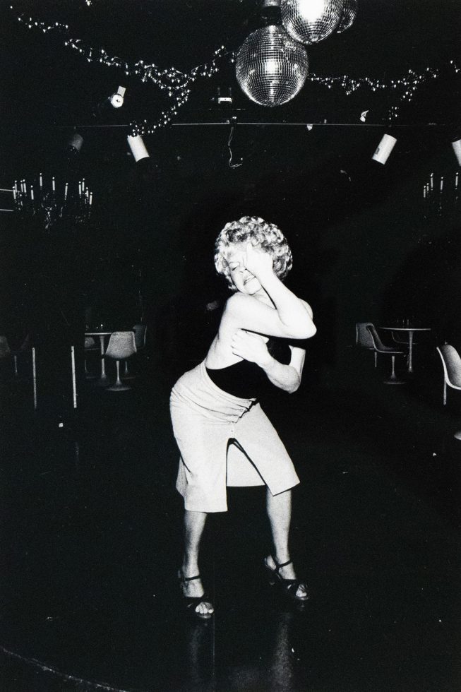 A woman with curly blonde hair and a dark top dances in a dimly lit room, with her hand raised to her face. Disco balls and string lights are visible overhead, suggesting a nightclub or performance venue.