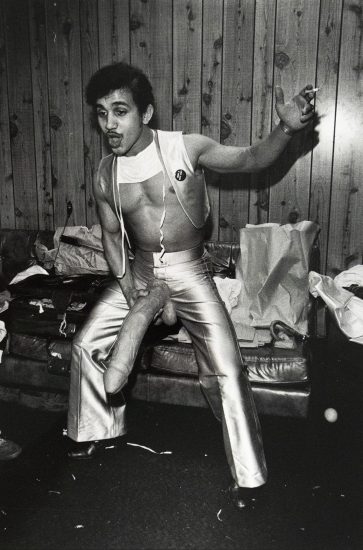 Black-and-white photograph of a man posing in front of a wood-paneled wall, wearing metallic pants and a cropped top. He holds a large dildo attached to his waist and has a cigarette in one hand, with a cluttered couch and paper bags in the background.