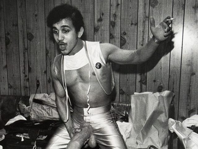 Black-and-white photograph of a man posing in front of a wood-paneled wall, wearing metallic pants and a cropped top. He holds a large dildo attached to his waist and has a cigarette in one hand, with a cluttered couch and paper bags in the background.