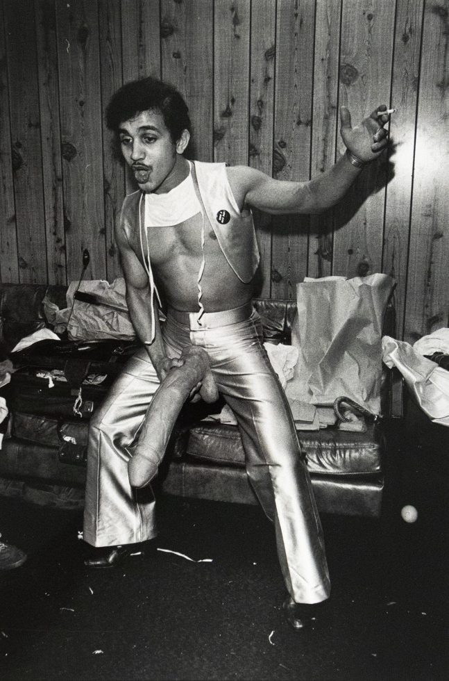 Black-and-white photograph of a man posing in front of a wood-paneled wall, wearing metallic pants and a cropped top. He holds a large dildo attached to his waist and has a cigarette in one hand, with a cluttered couch and paper bags in the background.