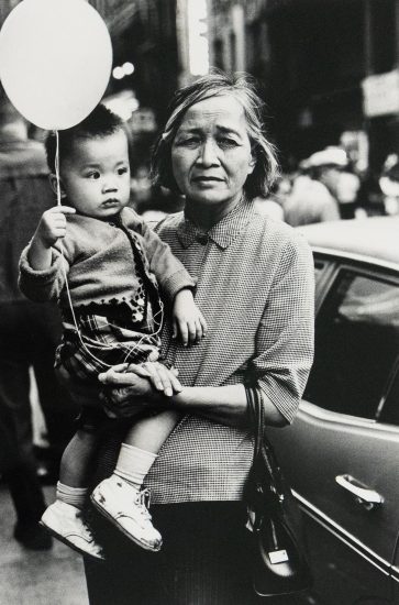 A black and white photograph shows a woman holding a child who clutches a balloon, both looking towards the right side of the frame. The scene is captured outdoors, with a vehicle visible behind them.