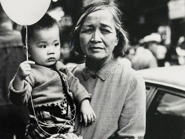 A black and white photograph shows a woman holding a child who clutches a balloon, both looking towards the right side of the frame. The scene is captured outdoors, with a vehicle visible behind them.