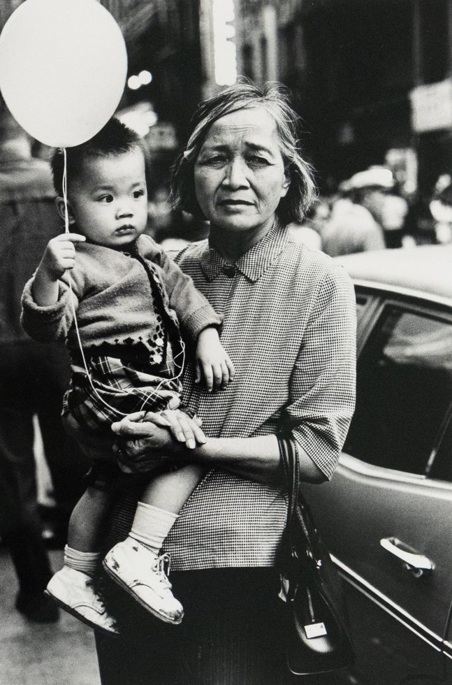A black and white photograph shows a woman holding a child who clutches a balloon, both looking towards the right side of the frame. The scene is captured outdoors, with a vehicle visible behind them.