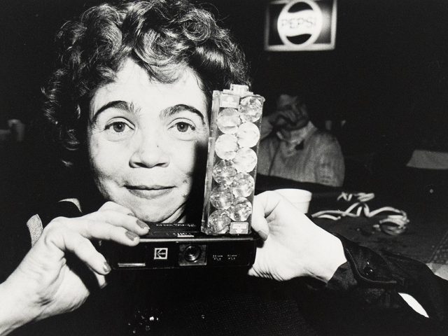 A woman holds a vintage camera decorated with clear gems at a dimly lit venue, with a Pepsi sign in the background.