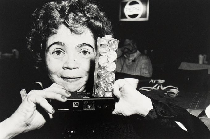 A woman holds a vintage camera decorated with clear gems at a dimly lit venue, with a Pepsi sign in the background.