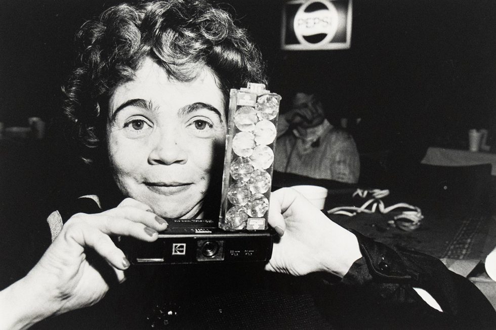 A woman holds a vintage camera decorated with clear gems at a dimly lit venue, with a Pepsi sign in the background.