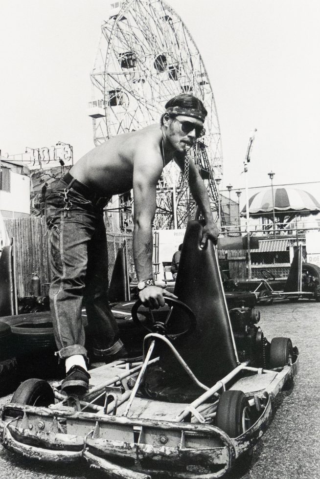 A shirtless man wearing a bandana and sunglasses stands over a go-kart, gripping its side, with a large Ferris wheel visible in the background.