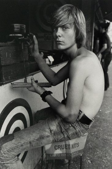 A black and white photograph captures a shirtless young man with shoulder-length blonde hair, sitting in profile while facing forward. He is positioned next to a game booth featuring targets and holds a set of darts, looking directly at the viewer with an intense expression.