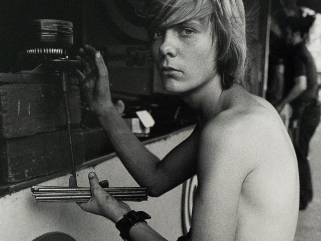A black and white photograph captures a shirtless young man with shoulder-length blonde hair, sitting in profile while facing forward. He is positioned next to a game booth featuring targets and holds a set of darts, looking directly at the viewer with an intense expression.