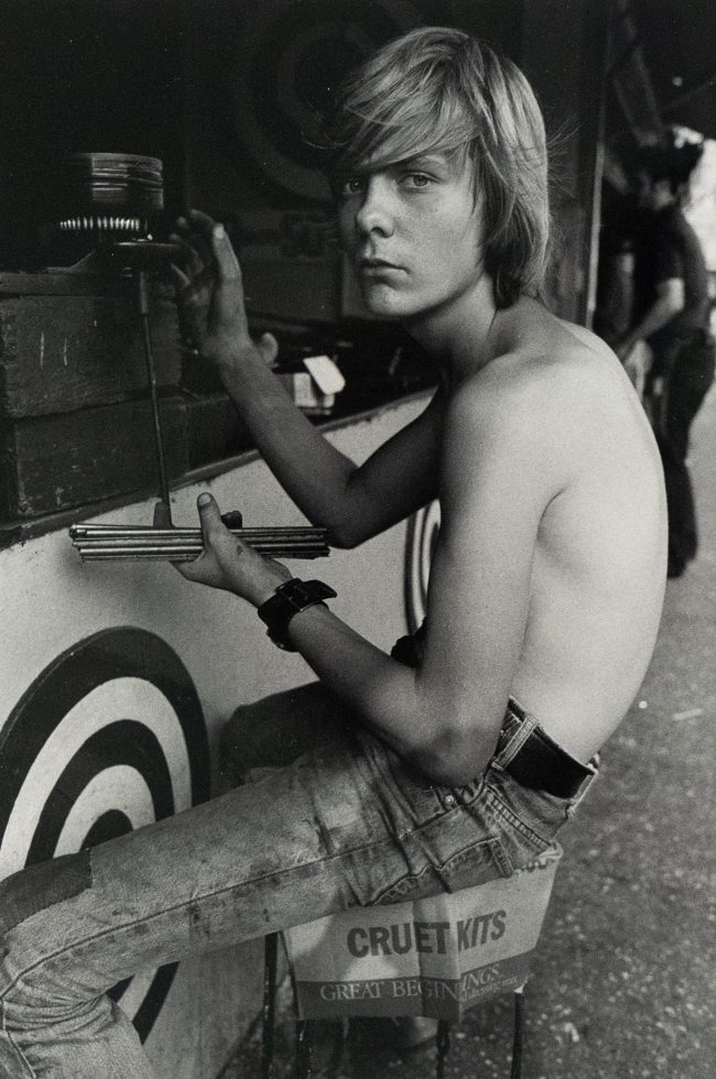 A black and white photograph captures a shirtless young man with shoulder-length blonde hair, sitting in profile while facing forward. He is positioned next to a game booth featuring targets and holds a set of darts, looking directly at the viewer with an intense expression.