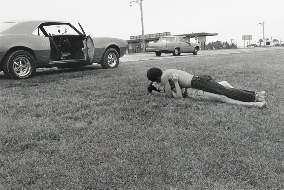 A couple lies embracing in a grassy field beside a highway, with a classic American muscle car parked nearby with its driver's side door open. In the background, other vehicles and structures are faintly visible under an overcast sky.