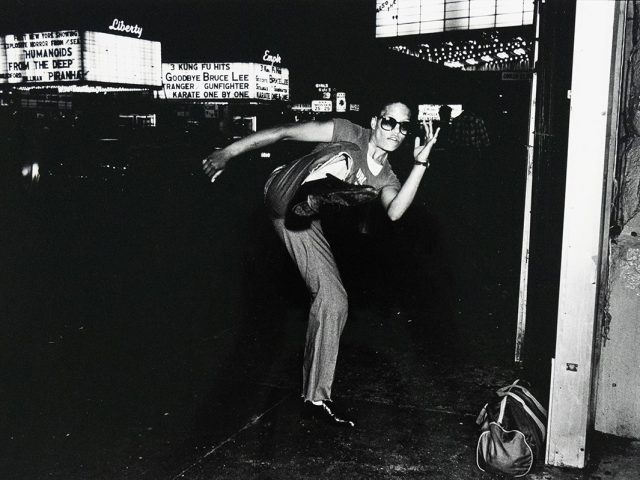 A black and white photograph captures a person in sunglasses striking a dynamic pose with one leg raised and arm extended, on a dark street at night. Brightly lit signs, including one that reads "Liberty," are visible in the background, and a bag rests on the ground to the right.
