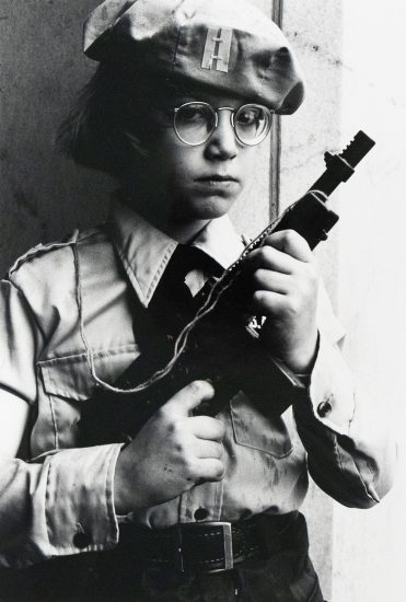 A person wearing a beret and round glasses holds a toy gun, looking directly at the viewer. The black and white image shows them from the waist up, dressed in a collared shirt with epaulets.