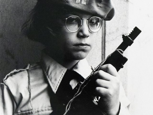 A person wearing a beret and round glasses holds a toy gun, looking directly at the viewer. The black and white image shows them from the waist up, dressed in a collared shirt with epaulets.