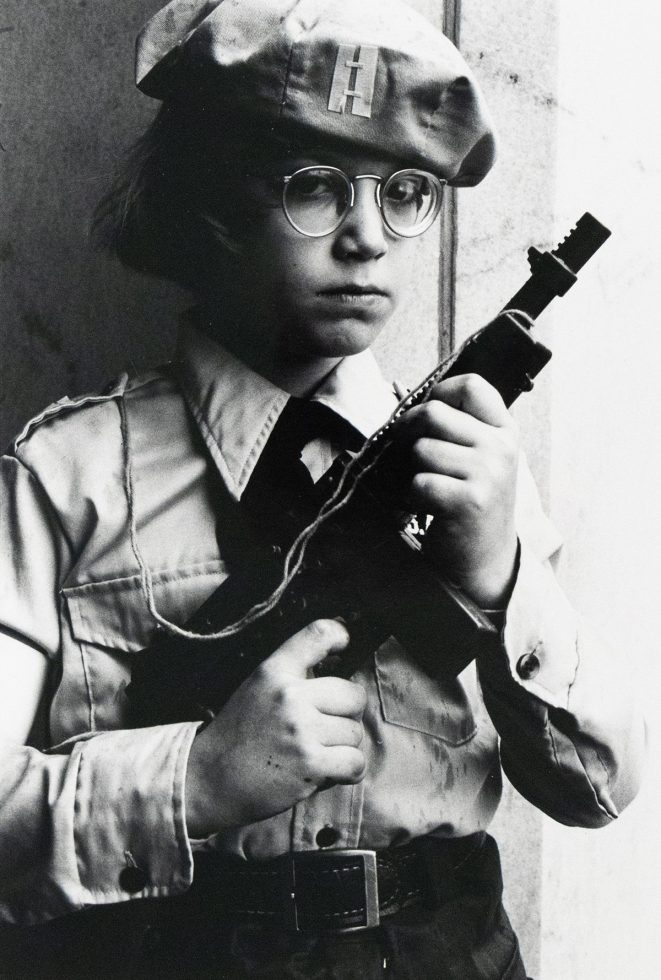 A person wearing a beret and round glasses holds a toy gun, looking directly at the viewer. The black and white image shows them from the waist up, dressed in a collared shirt with epaulets.